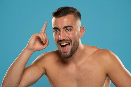 Young happy shirtless caucasian man 20s holding index finger up with great new idea isolated on blue...