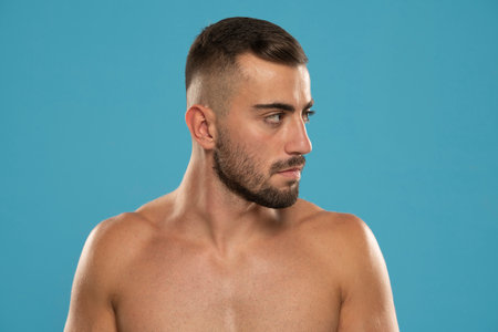 Handsome adult shirtless man withbeard and serious, looking aside confident standing against studio blue backgroundの写真素材