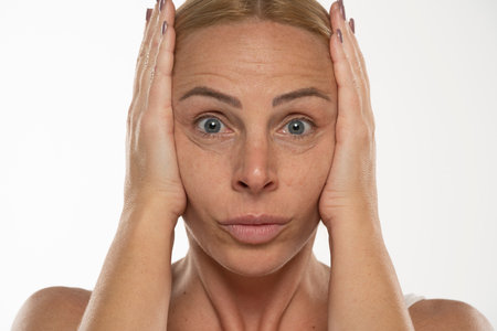 Middle aged blond woman tightens her face with her hands on a white studio backgroundの写真素材