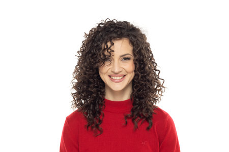 Portrait shot of beautiful young woman in red blouse with curly dark hair looking at camera with charming cute smile while posing against white studio backgroundの写真素材