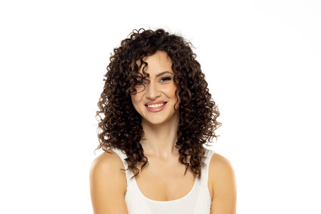 Portrait shot of beautiful young woman model with curly dark hair looking at camera with charming cute smile while posing against white studio backgroundの写真素材