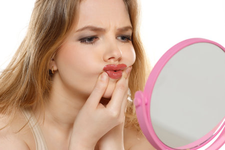 worried young blond woman in front of the mirror touches her mouth on a white studio backgroundの写真素材