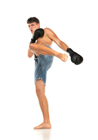 Full lenght shot of shirtless and barefooted young Caucasian male fighter raising one leg, ready to kick invisible enemy. Boxing, kickboxing, martial arts, sports, power on a white studio backgroundの写真素材