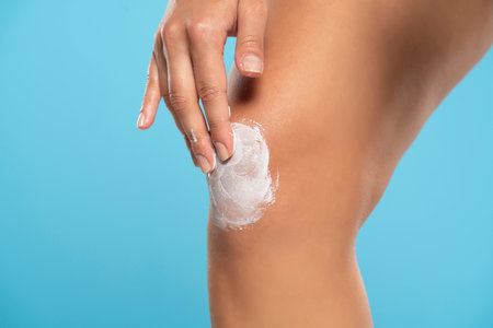 Cropped shot of young woman applying body cream on her knee on a blue studio backgroundの写真素材