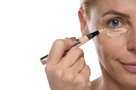 Middle aged senior woman applyes concealer under her eyes on white studio backgroundの写真素材