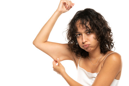 Unhappy etnic woman testing the flabby muscle under her arm pulling it down with her hand as she checks for muscle tone or weight gain on a white studio backgroundの写真素材