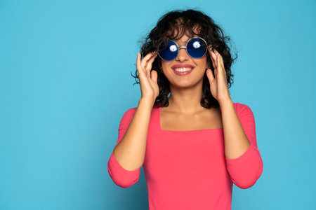 Young exotic smiling brunette woman in pink blouse posing with a sunglasses on a blue studio backgroundの写真素材