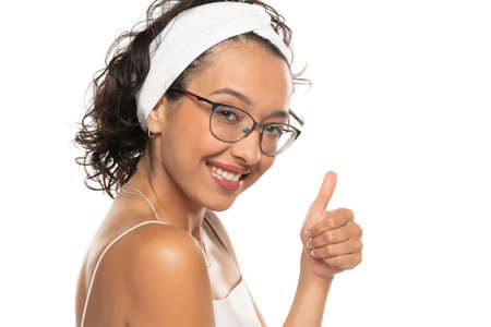 Young dark skinned smiling woman with makeup, headband and glasses posing on a white studio background. Thumbs up.の写真素材
