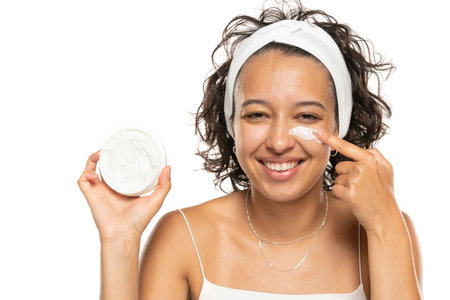 Young indian woman white shirt standing isolated on white studio background holding jar showing finger dipped in cream posing to camera smiling joyfulの写真素材