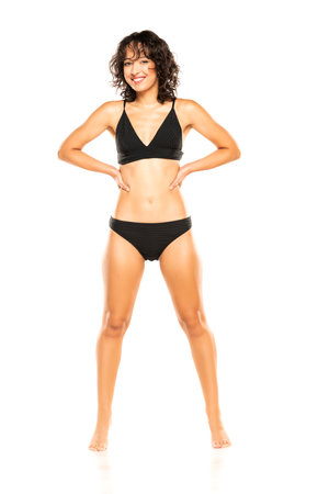 Young smiling exotic brunette woman in black bikini swimsuit posing on a white studio background. Front view.の写真素材