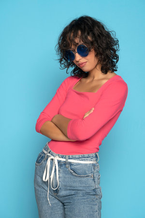 Young exotic brunette woman in pink blouse and rope belt jeans posing with a sunglasses on a blue studio backgroundの写真素材