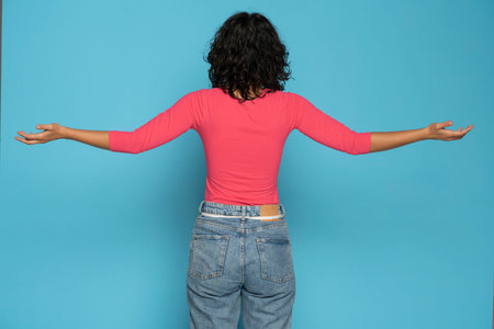 Rear view of a woman in jeands with wide open arms on a blue studio background