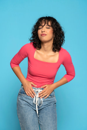 Young exotic woman having painful stomachache with hands holding pressing her lower abdomen. Medical or gynecological problems, healthcare concept on a blue studio backgroundの写真素材