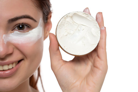Portrait of adorable, stylish model having white jar with  cream, applying under eyes, botox effect, fighting with dark circles, looking at camera isolated on white studio backgroundの写真素材