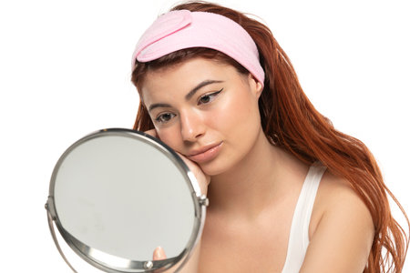 Young redhead beautiful woman with headband looks her self in mirror in a white studio backgroundの写真素材