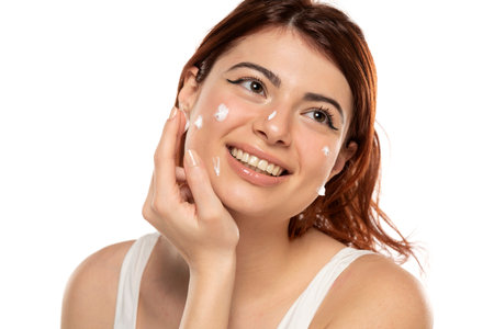 Beauty portrait image of pretty redhead smiling woman applying face cream isolated over white studio backgroundの写真素材