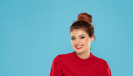 Cheerful gorgeous young woman wearing her ginger hair in bun smiling happily dressed in red blouse looking at camera with excited joyful smile on a blue studio backgroundの写真素材
