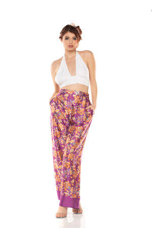 Fashion style studio portrait of beautiful young woman. Floral pattern wide leg pants and white sleevles shirt. Model standing and posing against white studio backgroundの写真素材