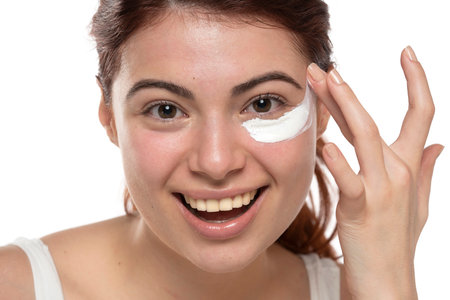 Beauty young smiling redhead woman applying cosmetic cream under her eye on a white studio backgroundの写真素材