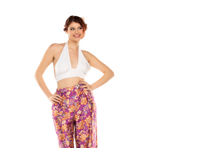 Fashion style studio portrait of beautiful young woman. Floral pattern wide leg pants and white sleevles shirt. Model standing and posing against white studio backgroundの写真素材