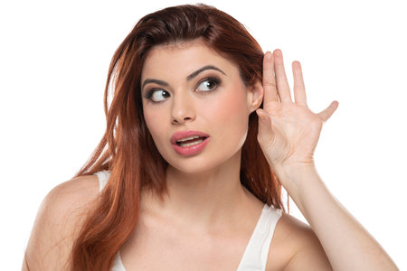 Redhead woman holds her hand near ear and listens carefully isolated on white studio backgroundの写真素材