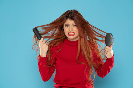 nervous young redhead woman with brush stuck in her messy hair on a blue studio background