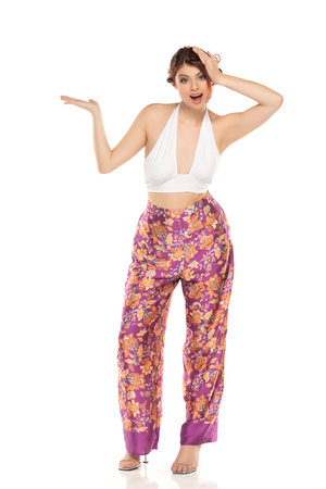 Smiling Excited Redhead Woman In Floral Pants And Sleevles Shirt Presenting Something On her Empty Palm On A White Studio Backgroundの写真素材