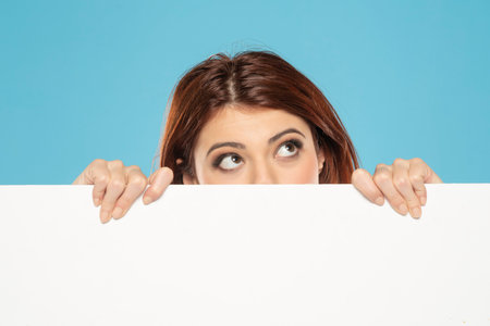 Banner of young thinking redhead woman peeking behind empty white canvas frame for text or advertising isolated on blue studio backgroundの写真素材
