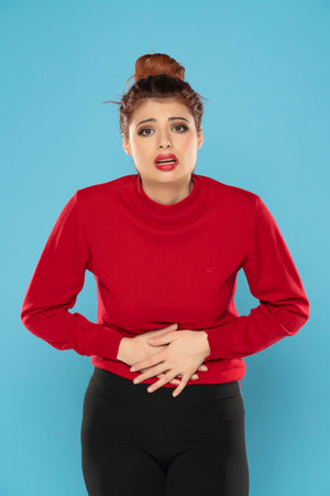 Redhead woman suffering from stomach pain and holding her hands in the lower abdomen on a blue studio backgroundの写真素材