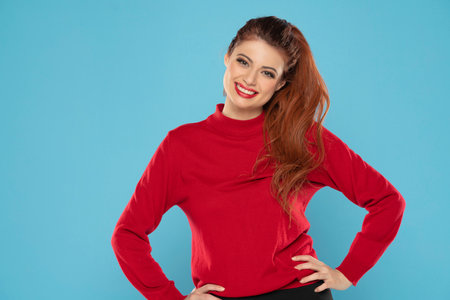 Cheerful gorgeous young woman wearing her ginger hair in ponytail smiling happily dressed in red blouse looking at camera with excited joyful smile on a blue studio backgroundの写真素材