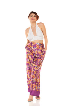 Fashion style studio portrait of beautiful young woman. Floral pattern wide leg pants and white sleevles shirt. Model standing and posing against white studio backgroundの写真素材