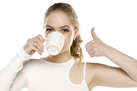 Beautiful young blond woman drinking coffee and showing thumb up on white studio backgroundの写真素材