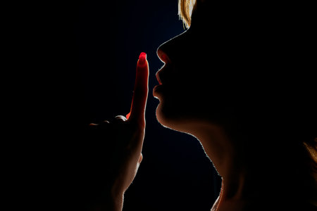 Silhouette of unknown woman , face in the shadow holding finger on her lips on a black studio backgroundの写真素材