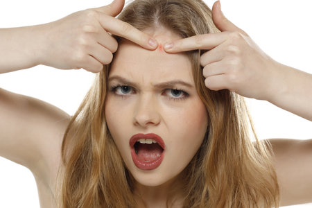 Nervous beautiful blonde woman squeeze pimples on her face on a white studio backgroundの写真素材