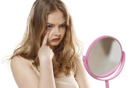 Young blond woman looking in the mirror nervous about the crack face skin and dark circle eyes on a white studio background.の写真素材