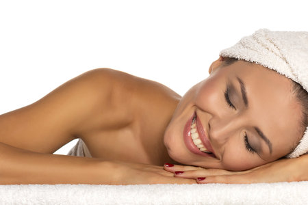Beauty portrait of a cheerful attractive half naked woman with a towel wrapped around her head with toothy smile and closed eyes lying on white studio backgroundの写真素材