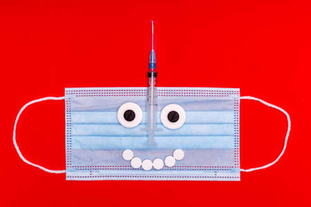 medical mask with a face made of pills and a syringe on a red backgroundの写真素材