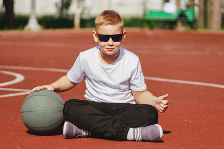 caucasian boy blond sits with a basketball ball on the sports groundの写真素材