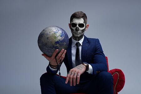 Satan holds the world in his hands. Planet earth in the hands of death. Man With Body Art Skeleton. Portrait of a skeleton in a suit looking at the camera.の写真素材