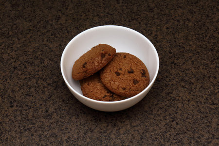 Oatmeal cookies with chocolate pieces in white bowl on the brown table