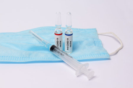 Two medicine ampule with anti covid-19 vaccine and syringe on protection mask on white background