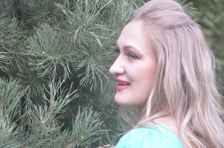 Beautiful young woman with blond hair and blue eyes in a turquoise blouse on the background of a green treeの写真素材