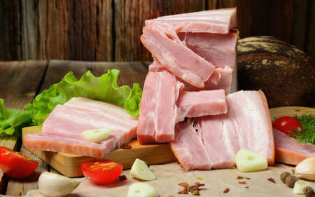 Smoked sliced pork belly, bacon, ham with herbs, tomatoes and black pepper on food paper. Wooden rustic background.Selective focus.Horizontal view.の写真素材