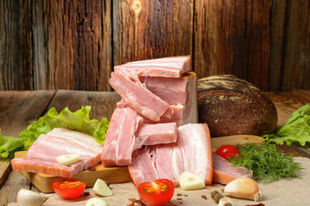 Smoked sliced pork belly, bacon, ham with herbs, tomatoes and black pepper on food paper. Wooden rustic background.Selective focus.Horizontal view.の写真素材