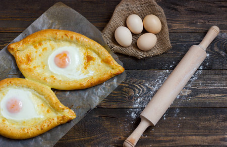 Khachapuri with eggs on sackcloth, flour, eggs and rolling pin on black table