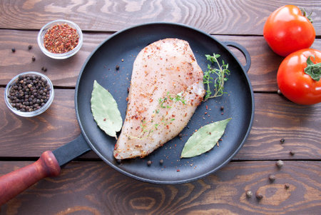 raw steak from chicken fillet on an iron black frying pan with herbs and spices, laurel leaf and branches of thyme near red ripe tomatoes and pepper peasの写真素材