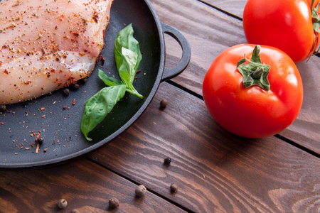 ripe red tomatoes on a wooden table next to a frying pan on which a fresh raw chicken fillet is sprinkled with herbs and spicesの写真素材