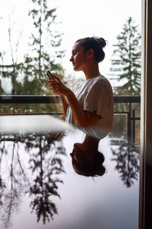 Close up of a woman profile with cellphone. Vertical shot, artistic reflection of a view from the window. Duality concept.の写真素材