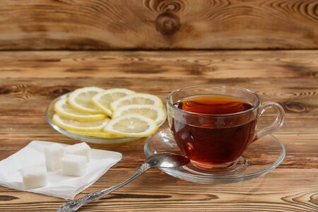 A glass cup of tea on a wooden table with sugar and slices of lemon in a plate.の写真素材