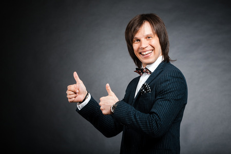 happy handsome man with long hair brunette and brown eyes in dark suit with stripes and bow tie and showing two thumb up sign looking forward at black backgroundの写真素材